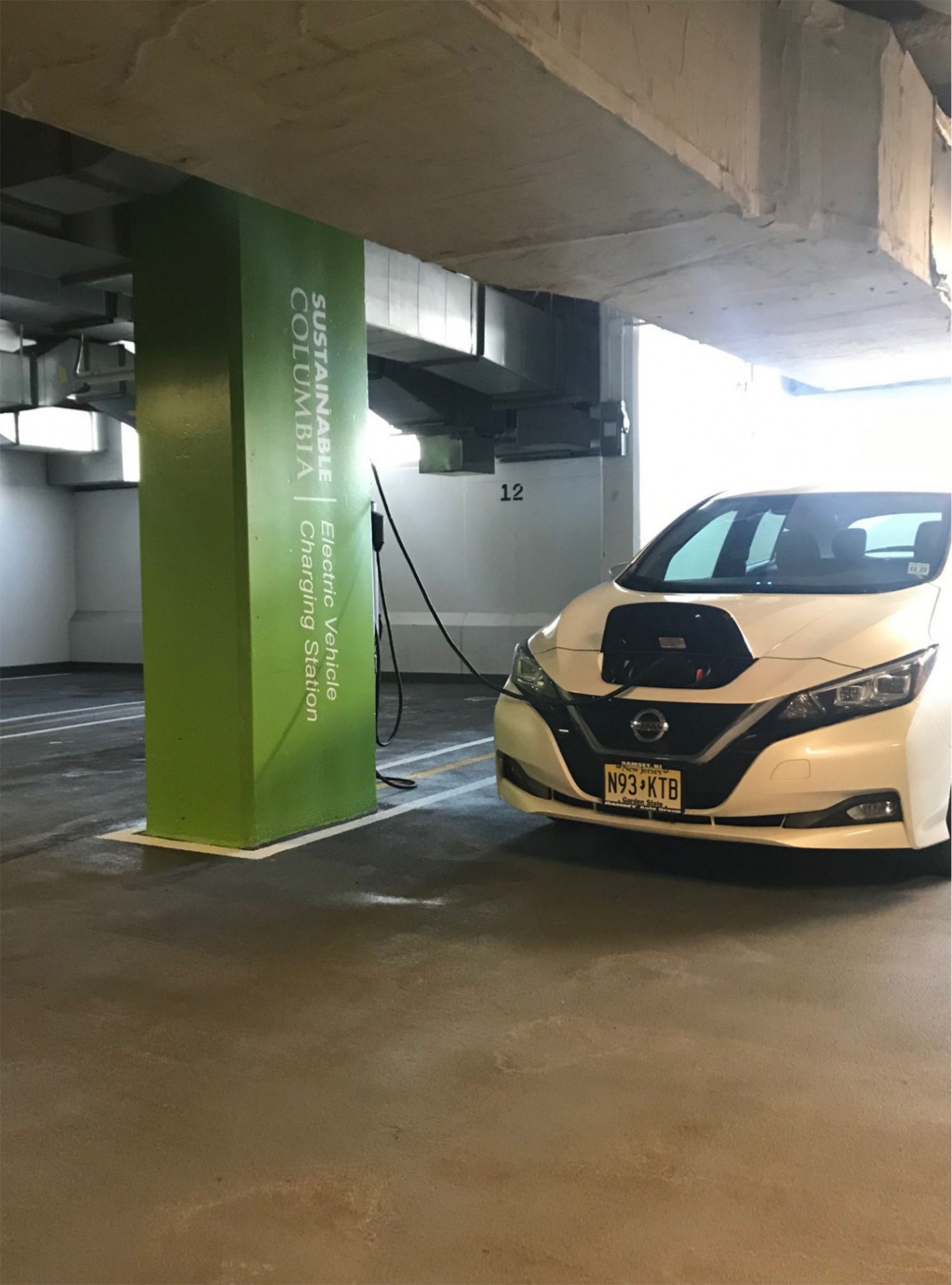 An electric car charging in the Columbia garage with the Sustainable Columbia green and logo on a pillar in the background.