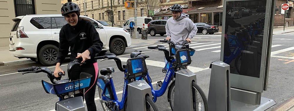 Two people wearing helmets and walking Citi Bikes to a dock at an intersection in Washington Heights.