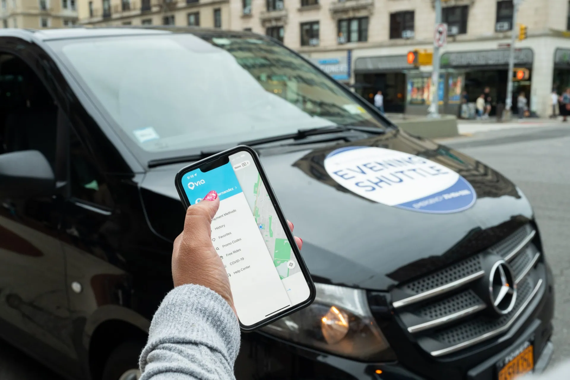 A person holds a smartphone with the Evening Shuttle app pulled up. They stand in front of a black van with the Evening Shuttle logo on the hood.